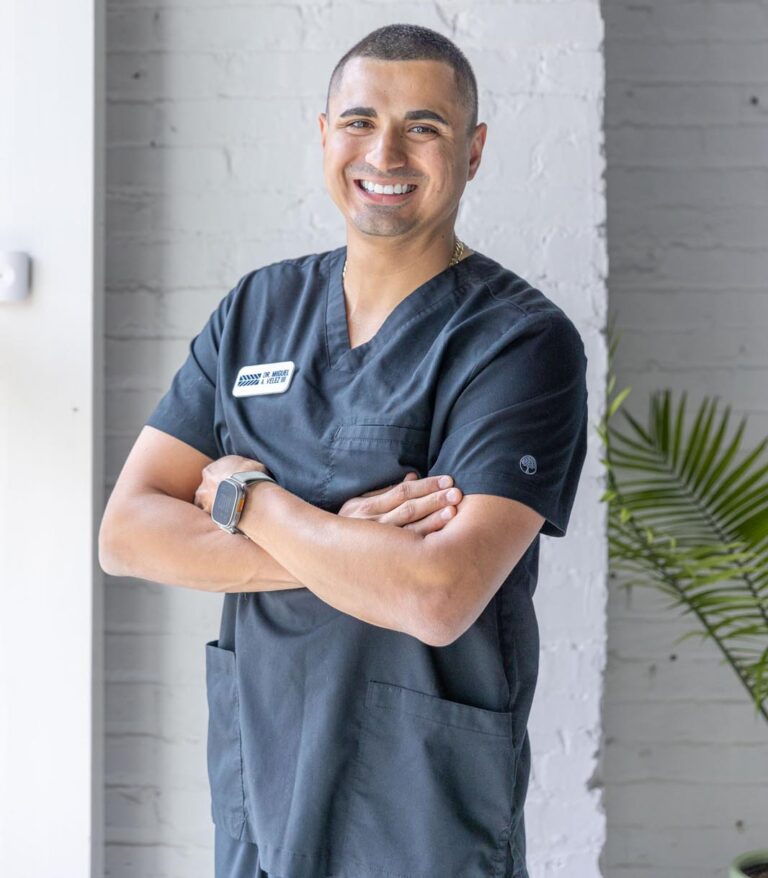 Dr. Miguel Velez hero shot in dark medical scrubs with a name tag and smartwatch, standing with arms crossed in front of a light-colored brick wall. The background includes a partial view of a green plant, suggesting a professional healthcare setting.