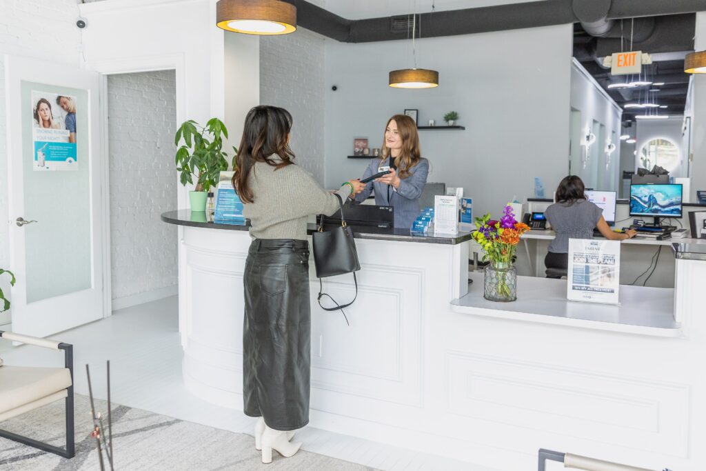 The reception area of a modern office or clinic featuring a curved white reception desk. A woman hands an item to a receptionist, while another person works on a computer behind the desk; decorative elements include colorful flowers and brochures, with plants and hanging lights enhancing the sleek design.