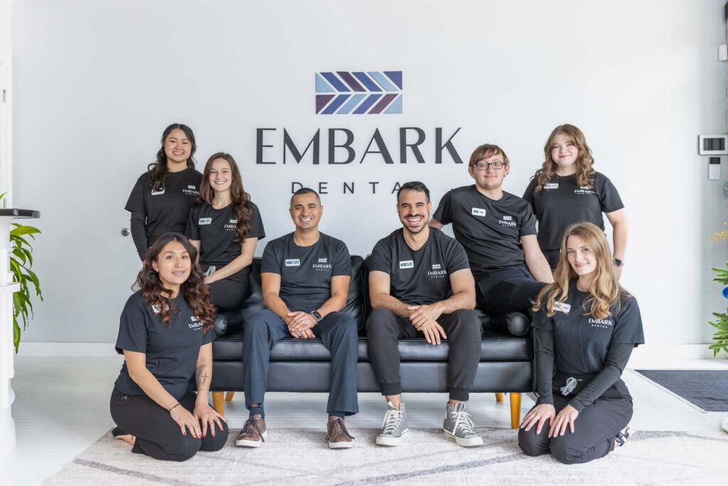 Group photo of eight individuals wearing black t-shirts with the logo 'EMBARK DENTAL.' They are posed in front of a wall displaying the 'EMBARK DENTAL' name, with three seated on a couch and five standing or kneeling around them, suggesting a professional team environment.