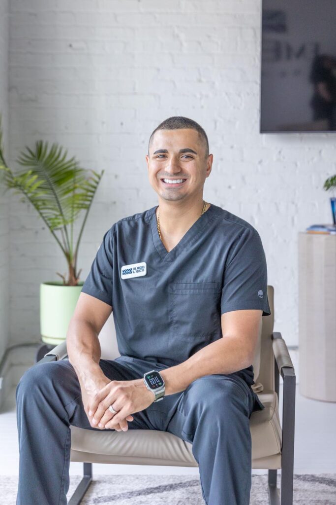 Dr. Miguel Velez sitting in a chair within a professional setting, wearing dark medical scrubs and an Apple Watch. The backdrop includes a white brick wall, a mounted television, and items such as a green potted plant and furniture, suggesting a medical office or consultation space.